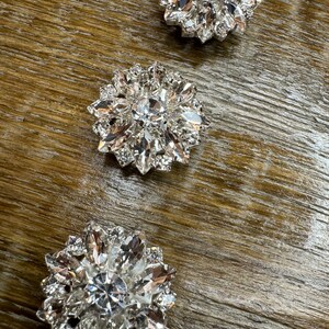 May include: Three silver-toned, rhinestone-encrusted embellishments in the shape of a flower. Each embellishment has a central rhinestone surrounded by smaller rhinestones.