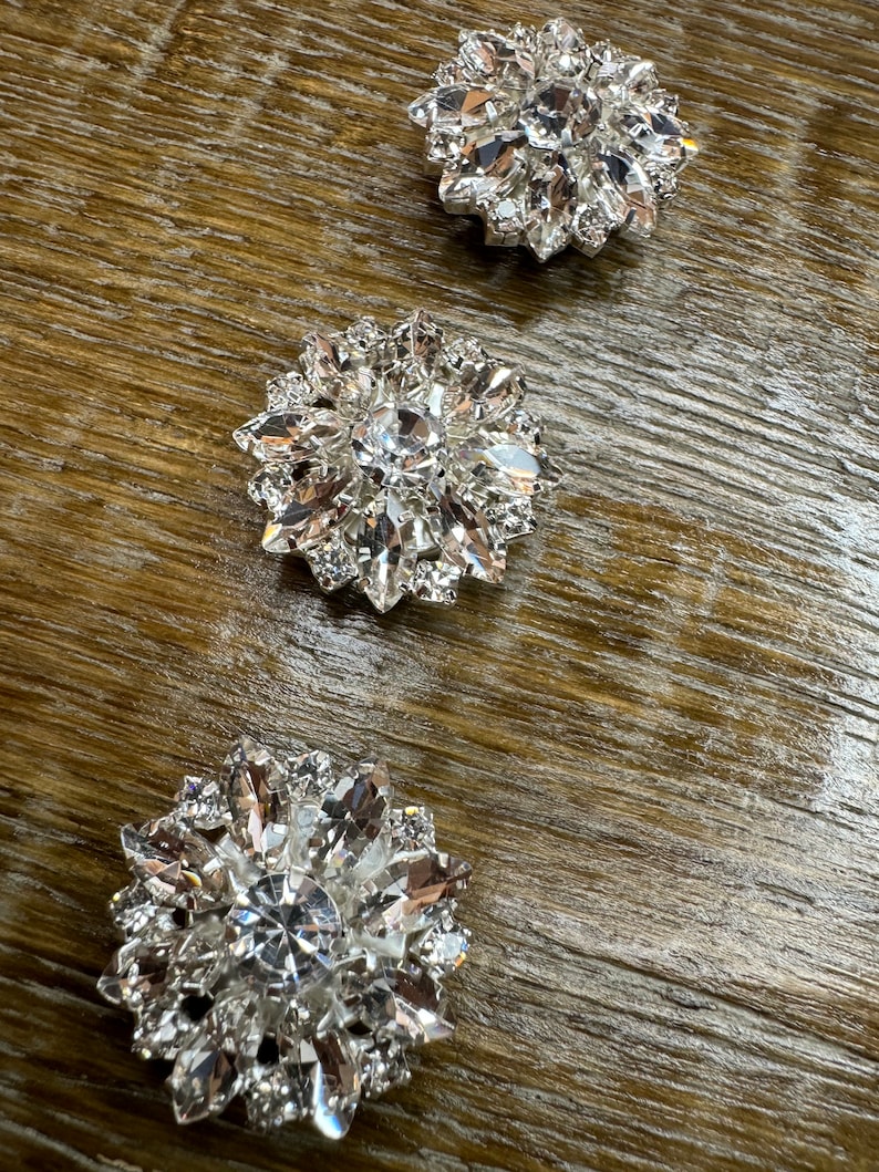 May include: Three silver-toned, rhinestone-encrusted embellishments in the shape of a flower. Each embellishment has a central rhinestone surrounded by smaller rhinestones.