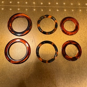 May include: Six round, tortoiseshell-patterned bangles in varying sizes. The bangles feature a mix of brown, black, and amber hues, with a glossy finish. The bangles are arranged on a light brown surface.
