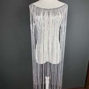 May include: A silver fringe garment featuring long, thin, metallic strands that drape from a wide, rounded neckline. The piece is constructed from numerous vertical, shimmering silver chains that create a fluid, layered effect over the torso.
