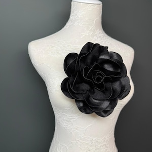 May include: A large, black fabric flower pinned to a white dress form. The flower has multiple layers of petals, creating a full, dimensional look. The dress form is covered in a delicate lace pattern, set against a grey background.