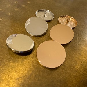 May include: Five round, flat buttons in two different colors. Two are silver, two are gold, and one is a silver button with a decorative design.