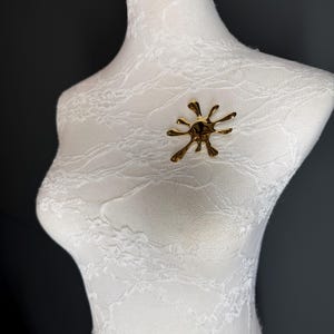 Puede incluir: Un broche abstracto dorado en forma de salpicadura o estallido de estrella. El broche est&aacute; en un maniqu&iacute; cubierto de encaje blanco.