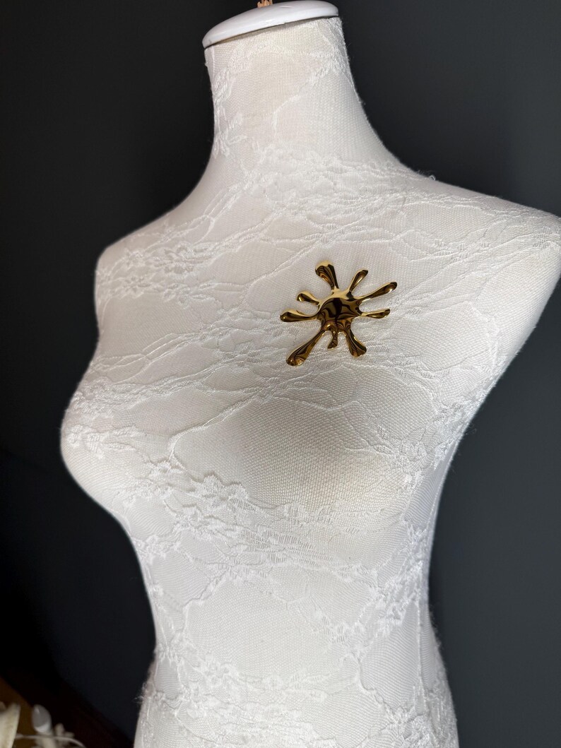 Puede incluir: Un broche abstracto dorado en forma de salpicadura o estallido de estrella. El broche est&aacute; en un maniqu&iacute; cubierto de encaje blanco.