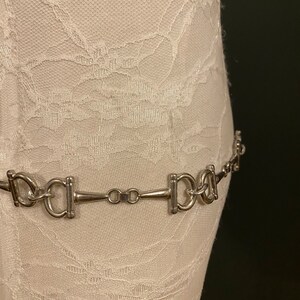 May include: A silver chain belt with a horse bit design. The belt is on a white fabric background.