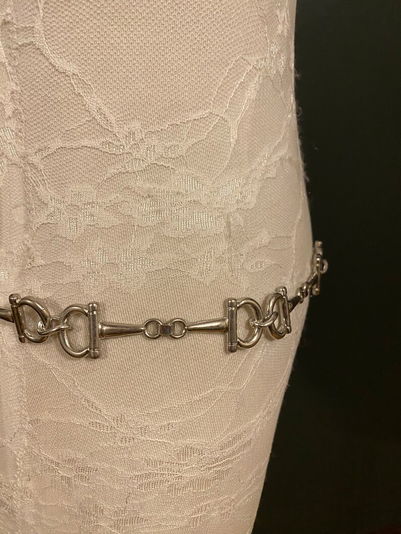 May include: A silver chain belt with a horse bit design. The belt is on a white fabric background.