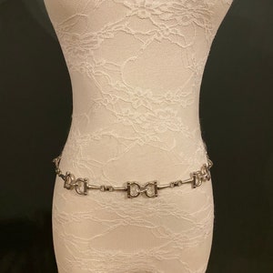 May include: A silver chain belt with a repeating pattern of interlocking D-shaped links. The belt is displayed on a white lace-covered mannequin.