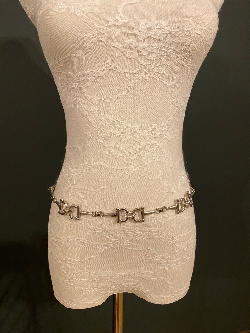 May include: A silver chain belt with a repeating pattern of interlocking D-shaped links. The belt is displayed on a white lace-covered mannequin.
