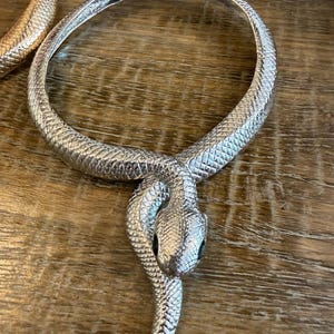 May include: A silver-toned metal choker necklace shaped like a coiled snake with a textured, scale-like surface pattern. The snake head rests at the centre, and the circular band features a hinged clasp mechanism at the top for opening and closing.