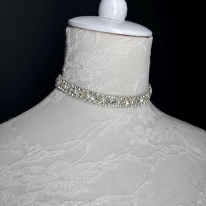 May include: A silver choker necklace with clear rhinestones is displayed on a white mannequin head. The necklace is made of small, round rhinestones and is designed to sit close to the neck. The mannequin is on a black background.