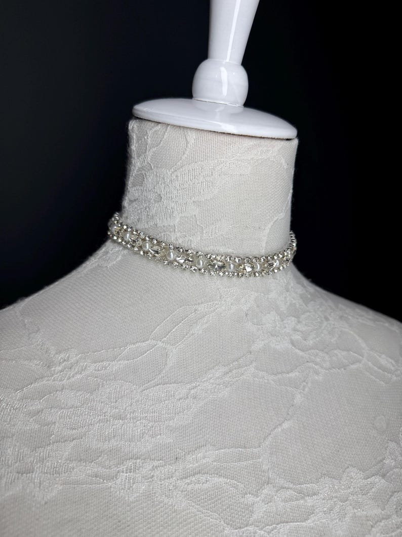 May include: A silver choker necklace with clear rhinestones is displayed on a white mannequin head. The necklace is made of small, round rhinestones and is designed to sit close to the neck. The mannequin is on a black background.