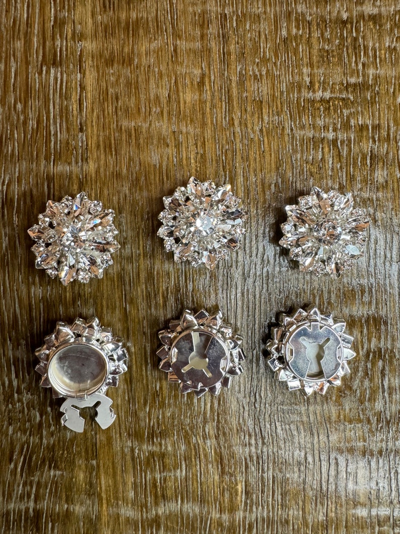 May include: Six silver-toned, rhinestone-encrusted embellishments. Three are shown with the rhinestones facing up, and three are shown with the back side facing up, revealing a metal clasp.