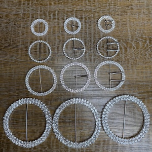 May include: A set of 10 silver-toned rhinestone round buckles of varying sizes. The buckles are arranged in three rows on a wooden surface.