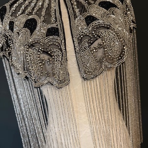 May include: A black and silver beaded cape with fringe. The cape features intricate patterns and a high collar. The silver beading is prominent, creating a shimmering effect. The fringe hangs down, adding movement to the design.