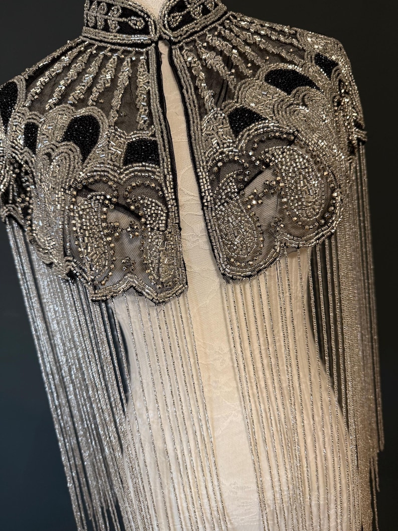 May include: A black and silver beaded cape with fringe. The cape features intricate patterns and a high collar. The silver beading is prominent, creating a shimmering effect. The fringe hangs down, adding movement to the design.