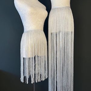 May include: Two white dress forms showcase silver fringe skirts. One skirt is knee-length, the other is floor-length. The fringe is made of thin, shimmering strands, creating a glamorous, flowing effect. The background is a solid black.