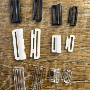 May include: A collection of twelve plastic strap adjusters and sliders arranged in three horizontal rows by color. The top row features four black pieces, the middle row has four white pieces, and the bottom row contains four clear pieces.