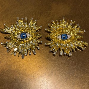 Könnte beinhalten: Ein Paar goldene Ohrringe mit einem blauen Augenmotiv und klaren Strasssteinen. Die Ohrringe sind sonnenstrahlenförmig.
