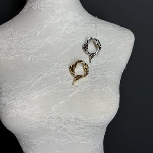 May include: Two abstract metal brooches, one gold and one silver, are displayed on a white mannequin. The brooches are shaped like a circle with an irregular, organic design.