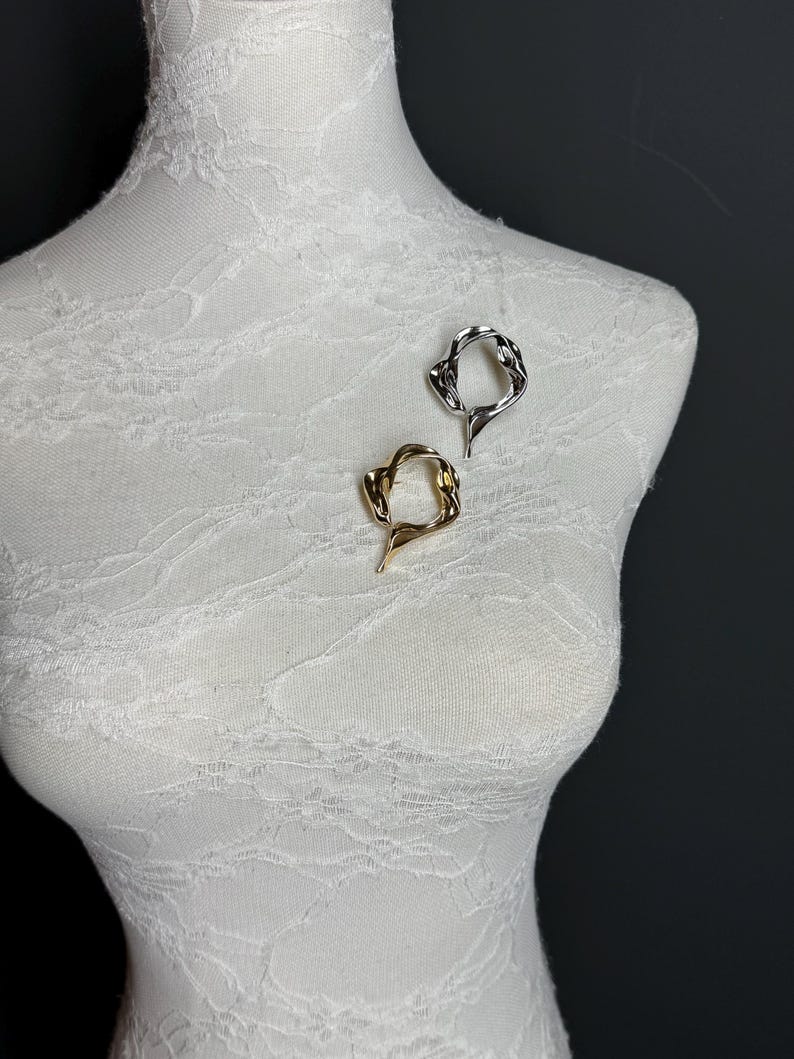 May include: Two abstract metal brooches, one gold and one silver, are displayed on a white mannequin. The brooches are shaped like a circle with an irregular, organic design.