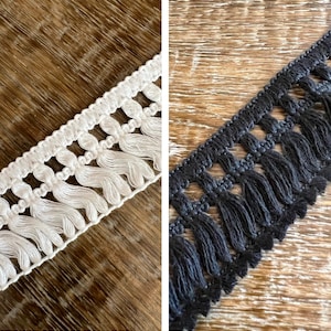 Könnte beinhalten: Zwei Muster von Fransenbesatz, eines weiß und eines schwarz. Beide Besätze haben ein sich wiederholendes Muster aus kleinen Schlaufen und Quasten.