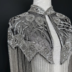 May include: A silver beaded capelet with a high collar and fringe. The capelet is made of black mesh and is heavily embellished with silver beads in floral and geometric patterns. The fringe hangs from the shoulders.