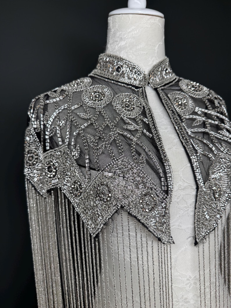 May include: A silver beaded capelet with a high collar and fringe. The capelet is made of black mesh and is heavily embellished with silver beads in floral and geometric patterns. The fringe hangs from the shoulders.