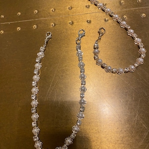 Puede incluir: Tres cadenas plateadas con detalles de perlas blancas y strass. Las cadenas son de diferentes longitudes y se muestran sobre una superficie dorada.