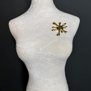 Puede incluir: Un broche de metal de color dorado en forma de salpicadura o goteo. El broche est&aacute; en un maniqu&iacute; blanco.