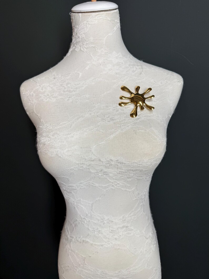 Puede incluir: Un broche de metal de color dorado en forma de salpicadura o goteo. El broche est&aacute; en un maniqu&iacute; blanco.