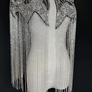May include: Silver beaded cape with detailed embellishments and long fringe. The cape has a geometric design around the neckline and shoulders. The fringe hangs down, creating a dramatic look. The cape is displayed on a white mannequin.