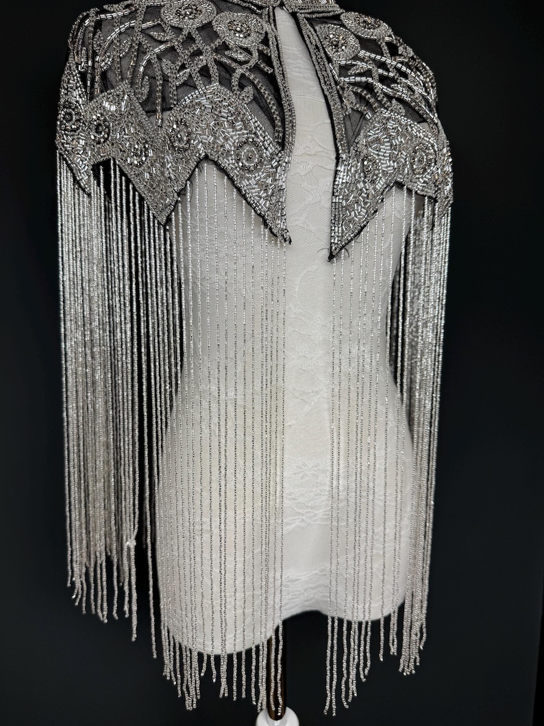 May include: Silver beaded cape with detailed embellishments and long fringe. The cape has a geometric design around the neckline and shoulders. The fringe hangs down, creating a dramatic look. The cape is displayed on a white mannequin.
