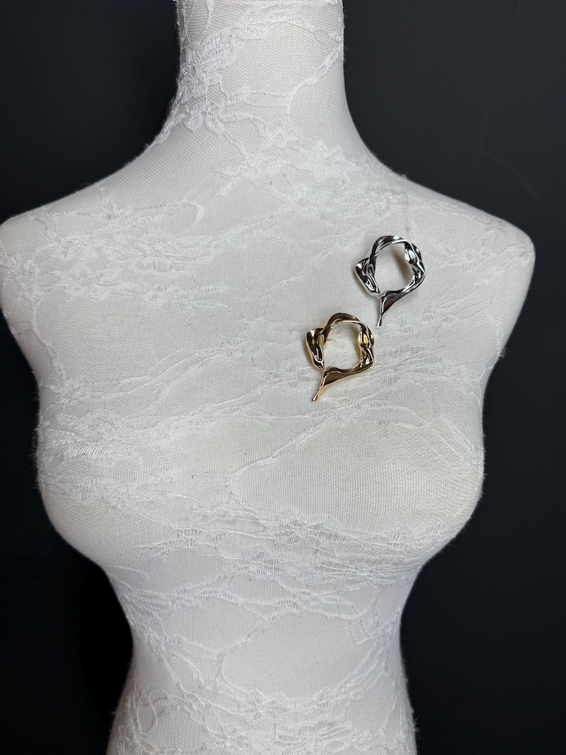 May include: Two abstract metal rings, one gold and one silver, are displayed on a white mannequin. The rings are both open and have a unique, organic shape.