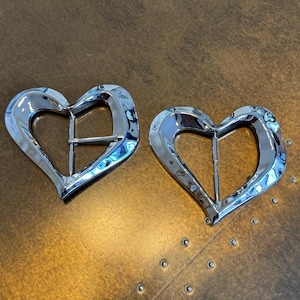 May include: Two heart-shaped belt buckles with a polished silver finish. Each buckle features a rectangular bar. The buckles are presented on a brown surface.