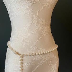 May include: A white pearl belt with a gold clasp. The belt is on a white lace mannequin.