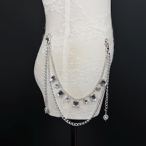 May include: Silver chain belt with multiple strands and clear crystal embellishments. The belt has two lobster claw clasps and a dangling crystal charm. The belt is displayed on a white mannequin. The crystals are rectangular and round.