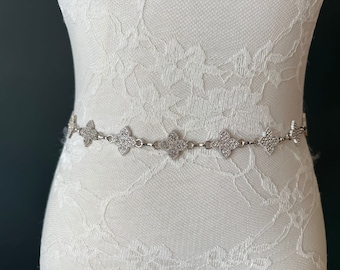 May include: A silver and clear crystal bridal belt with a delicate chain and a repeating pattern of four-leaf clover shapes.