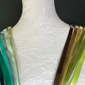 Puede incluir: Un conjunto de 10 cintas de satén de diferentes colores en tonos de verde, marrón y amarillo. Las cintas están dispuestas en forma de arcoíris sobre un maniquí blanco.