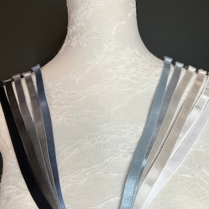 Puede incluir: Un conjunto de 10 tirantes de sujetador en varios tonos de gris, negro y blanco. Los tirantes están dispuestos en forma de abanico en un maniquí blanco.