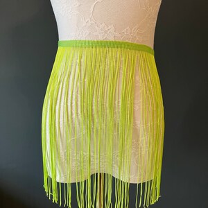 May include: A neon green fringe skirt displayed on a mannequin. The skirt has a woven waistband and long, vertical strands of fringe. The mannequin is set against a dark grey background.