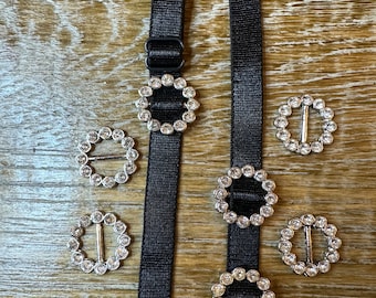 May include: Two black straps with silver metal clasps adorned with clear rhinestones. The straps are on a wooden surface with five additional clasps.