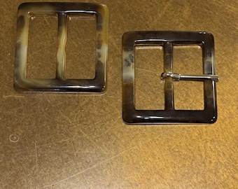 Puede incluir: Dos hebillas de cinturón cuadradas con un patrón marrón y beige veteado. Una hebilla tiene una barra de metal plateado. Las hebillas tienen un acabado brillante y una forma rectangular con dos barras interiores.