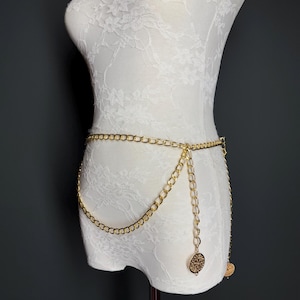 May include: A gold chain belt with two dangling circular charms. The belt is worn on a white lace mannequin.