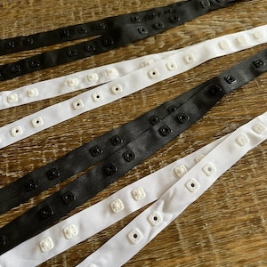 Può includere: Due cinturini elastici bianchi e due neri con bottoni di plastica bianchi e neri. I cinturini sono disposti su una superficie di legno.