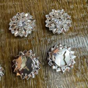 May include: Four silver-toned, round, decorative buttons with a clear crystal center. The buttons have a starburst design with pointed edges. Two buttons show the front side with crystals, and two buttons show the back side with a metal loop.