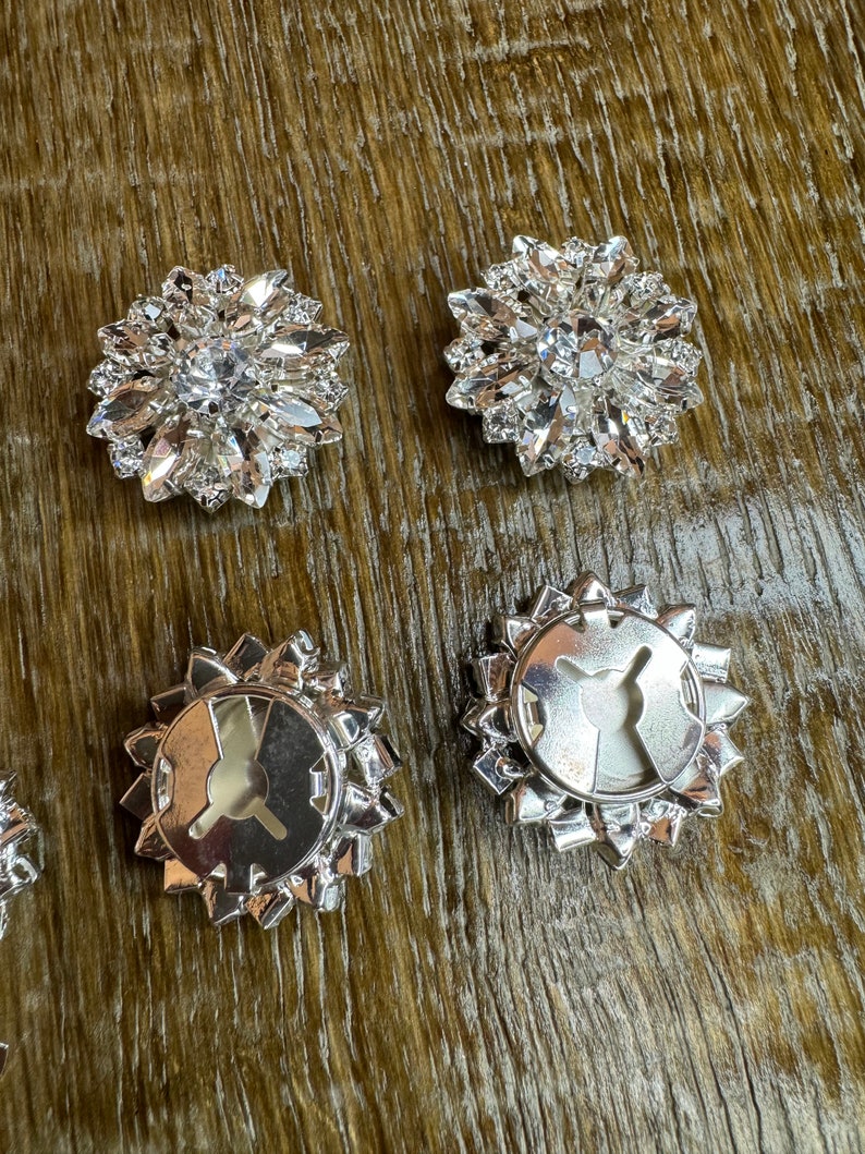 May include: Four silver-toned, round, decorative buttons with a clear crystal center. The buttons have a starburst design with pointed edges. Two buttons show the front side with crystals, and two buttons show the back side with a metal loop.