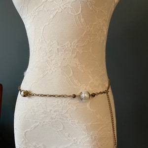Könnte beinhalten: Ein goldener Kettengürtel mit weißen Perlenakzenten. Der Gürtel befindet sich an einem weißen Spitzen-Mannequin.
