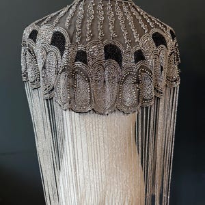 May include: A beaded cape with a high collar and fringe. The cape features intricate silver and black beadwork in a decorative pattern. Long, silver fringe hangs from the bottom edge, creating a flowing effect. The cape is displayed on a mannequin.