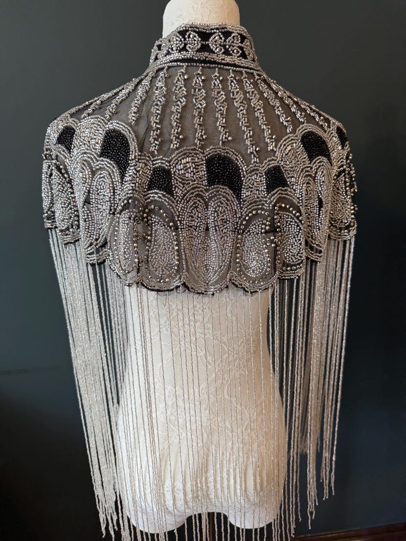 May include: A beaded cape with a high collar and fringe. The cape features intricate silver and black beadwork in a decorative pattern. Long, silver fringe hangs from the bottom edge, creating a flowing effect. The cape is displayed on a mannequin.