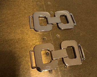 May include: Two silver metal belt buckle closures with a square shape and a connecting chain link. The buckles are on a gold surface.